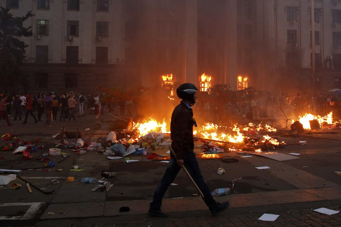 A protester wearing a Ukrainian flag walks past a burning tent camp and a fire in the trade union building in Odessa