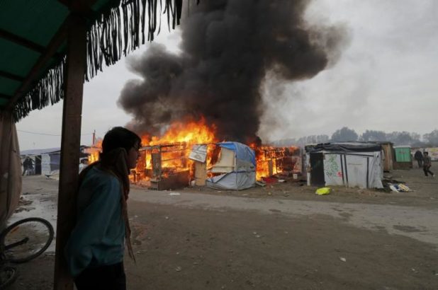 A migrant stands near a burning makeshift shelters in the "Jungle" on the third day of their evacuation, as part of the dismantlement of the camp in Calais